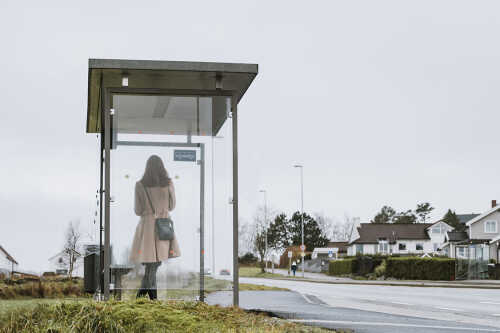 Bilde av dame i kåpe med veske som står og venter på buss i buss-skur.